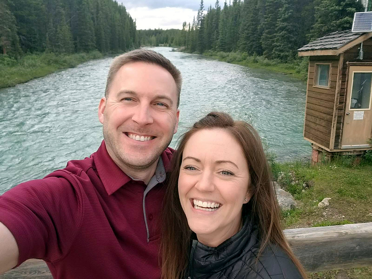 Matt and his wife smiling together on a bridge over a clear river, with evergreen forest behind them.